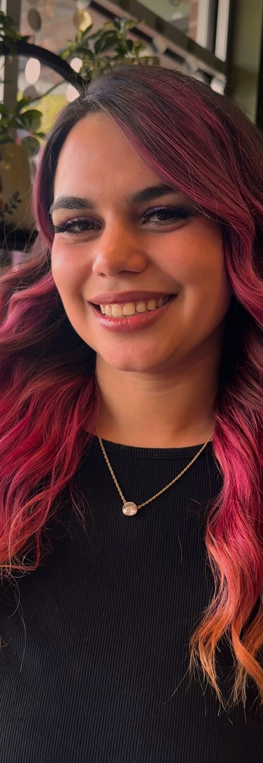 Sophie, smiling, with long magenta-to-copper waves, in a black top in front of a green wall and plants